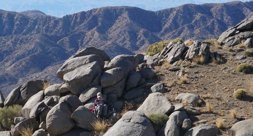 A person sits on a boulder with a mountain range in the background. They appear to be journaling. 